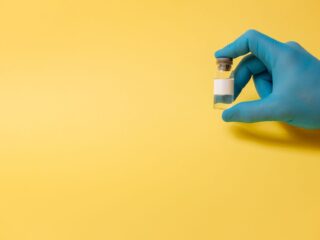 A hand in a blue glove holding a small vial against a yellow background